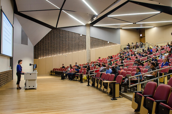 Teacher standing before projector in tiered lecture center half filled with students